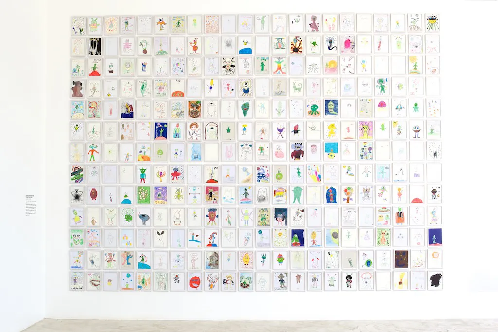 A white gallery wall hung with hundreds of alien drawings in a tight grid.