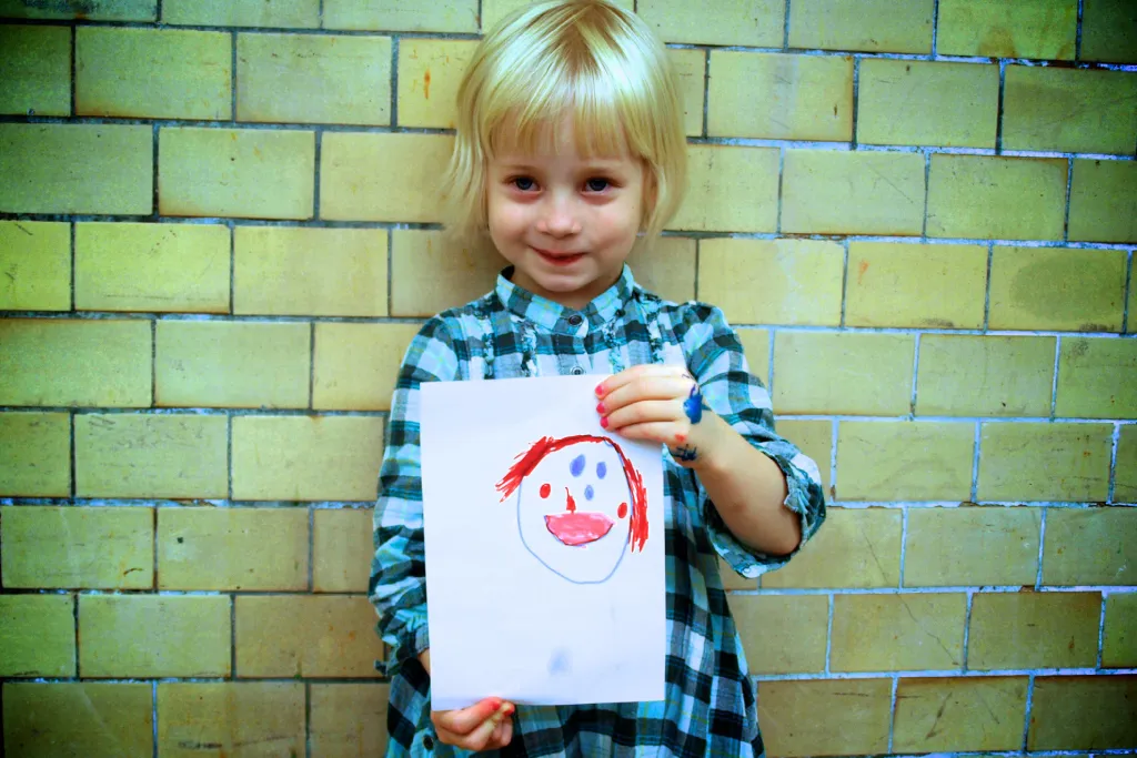 A young child holding a drawing of a face in front of a tiled wall.