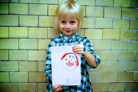 A young child holding a drawing of a face in front of a tiled wall.