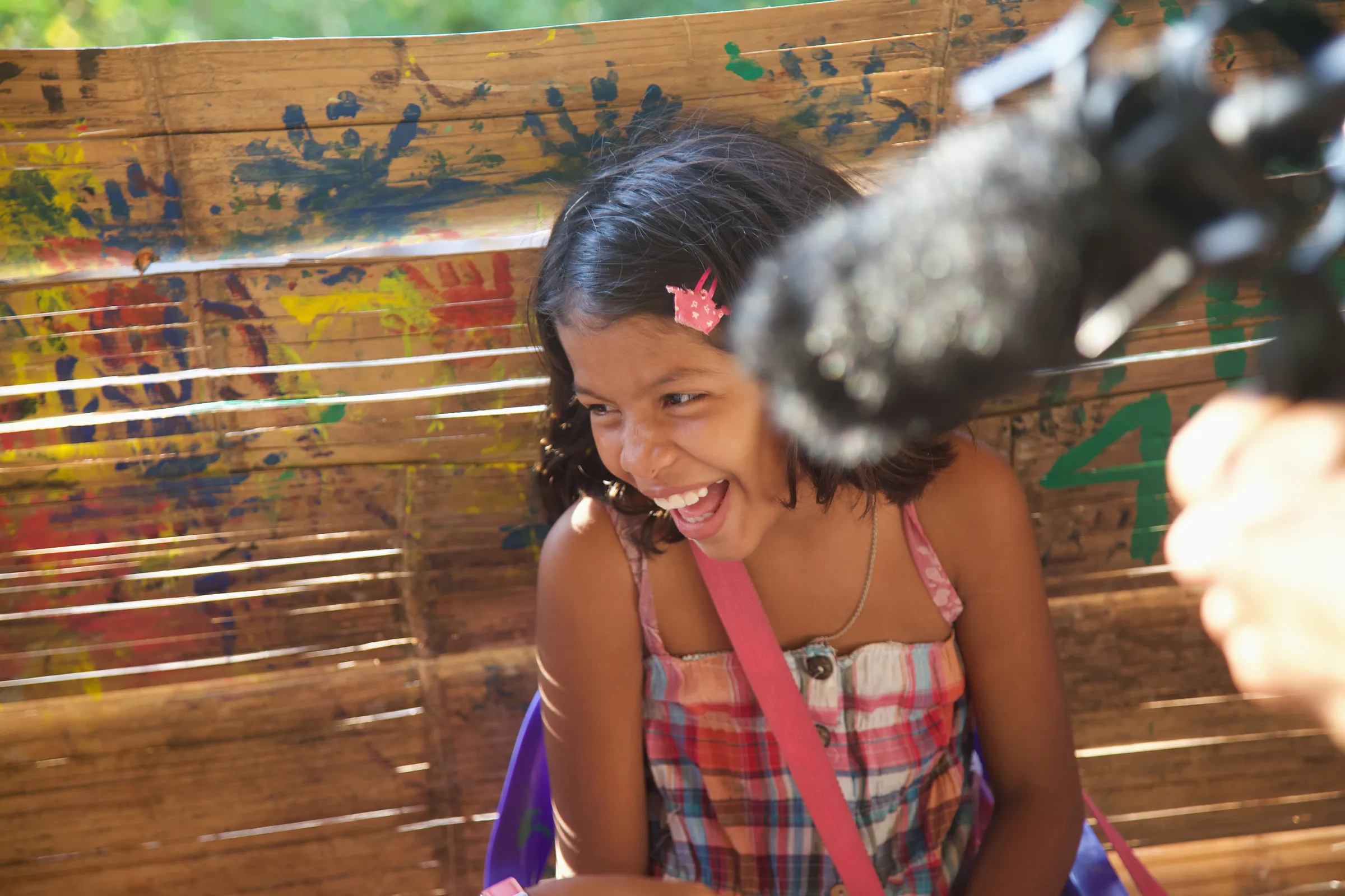 A child laughing beside a boom microphone, with painted handprints on the wall behind.