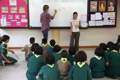 Two adults at a whiteboard in front of children seated on the floor.