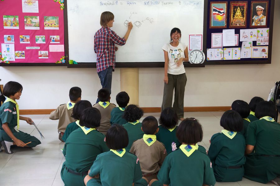 Two adults at a whiteboard in front of children seated on the floor.