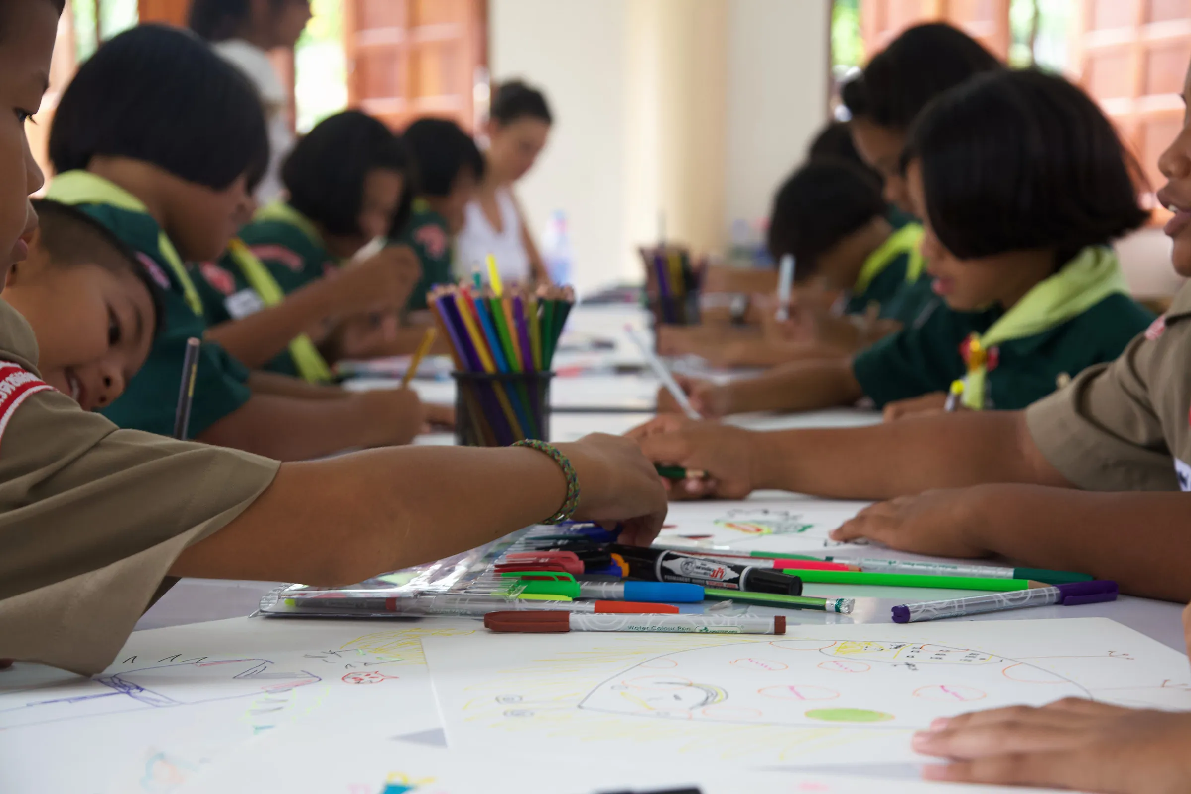 Children drawing with felt pens along a long table.