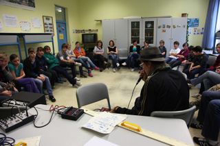 A man in a hat sings into a microphone at a loop machine, students seated in a circle. Brandenburg, Germany, 2010.