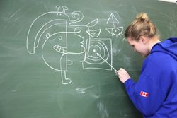 A girl in a blue hoodie adds to a large chalk drawing on a blackboard. Brandenburg, Germany, 2010.