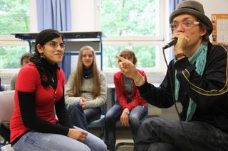 A man in a hat sings into a microphone and points at a student. Brandenburg, Germany, 2010.