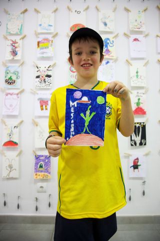 A boy in a yellow shirt holds up his drawing of a green alien on blue. Barcelona, Spain, 2010.