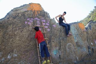 A boy climbs a wooden ladder up a boulder covered in chalk drawings and a painted sun. The Ideal Place, 2011.