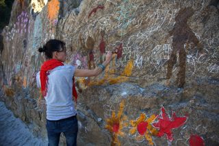 A woman in a red scarf paints on a rock covered in chalk lines and red stars. The Ideal Place, 2011.