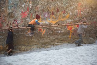 Children carry a long bamboo pole past a painted rock face. The Ideal Place, 2011.