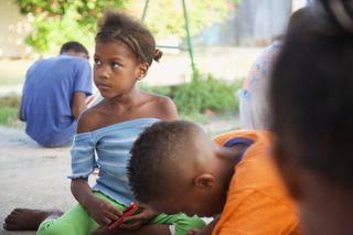 A girl in a blue top watches a boy in orange drawing on the ground. Barú, Colombia, 2011.