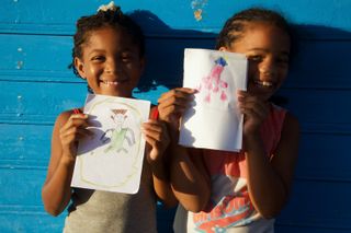 Two girls hold up their alien drawings against a blue wooden wall. Barú, Colombia, 2011.