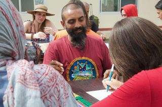 A bearded man in a red shirt draws at a table with others around him. Rishikesh, India, 2012.