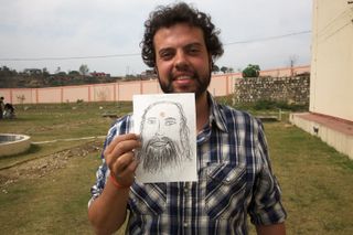 A curly-haired man holds up a drawing of a bearded face. Rishikesh, India, 2012.