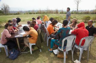 Rows of adults drawing at outdoor tables on a wide lawn. Rishikesh, India, 2012.