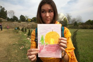 A woman in an orange scarf holds up a drawing of a green alien and a yellow sun. Rishikesh, India, 2012.