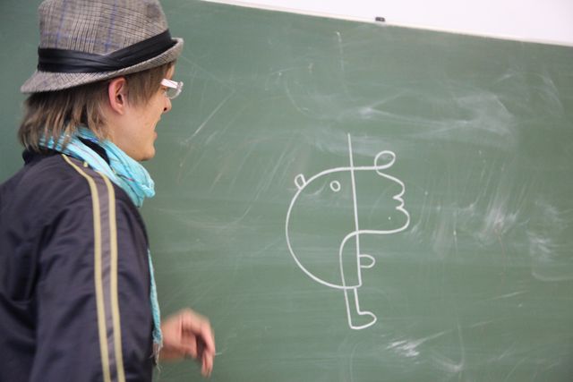 Ole Ukena in a hat and glasses, turning from a chalkboard with a line drawing of a figure.