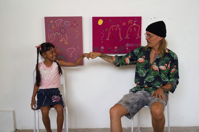 Ole Ukena and a girl sitting on two chairs, fists touching, under two pink embroidered panels.