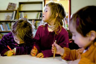 Three small children draw at a white table in front of bookshelves. Spain, 2012.