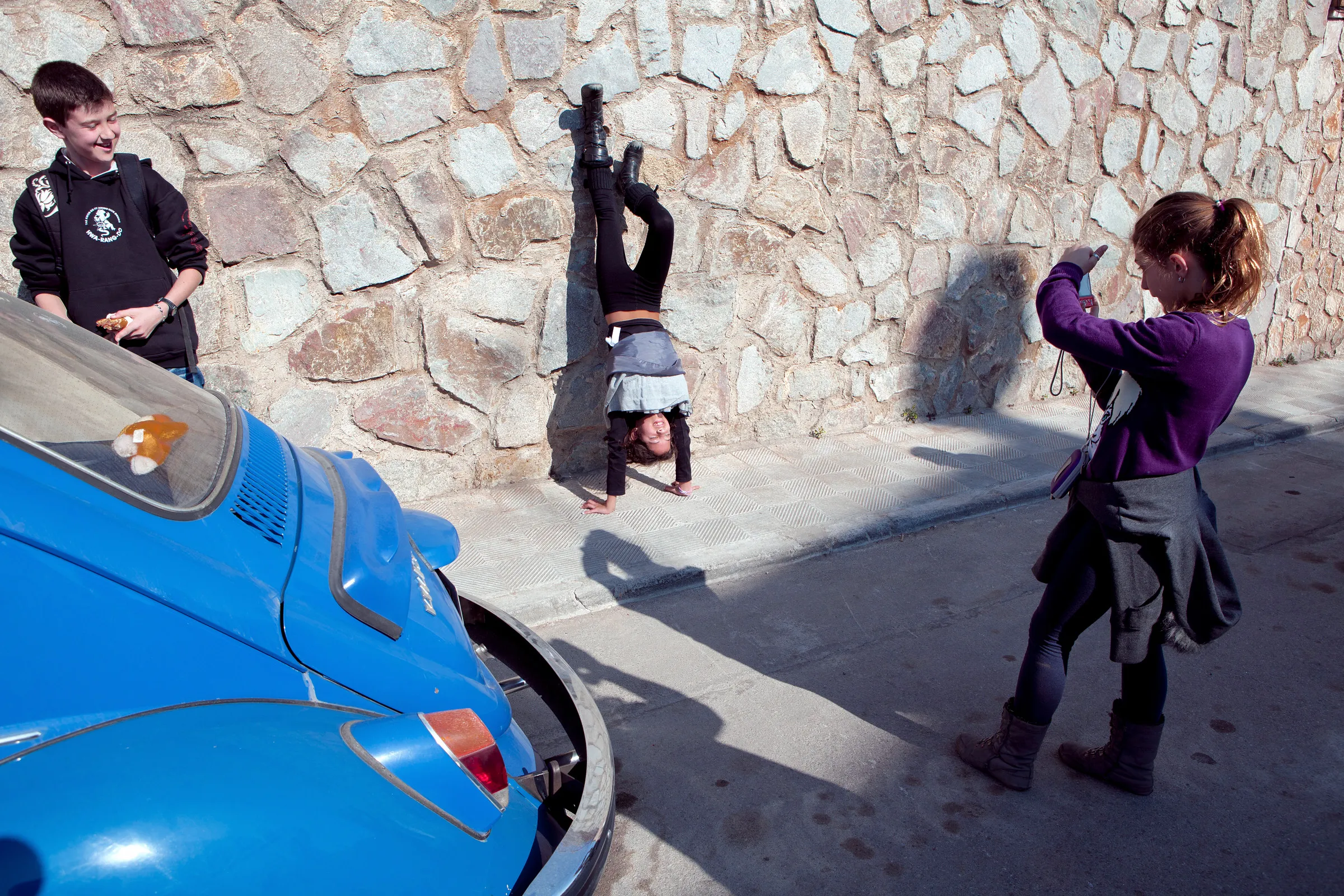 A child photographing another child doing a handstand against a stone wall.