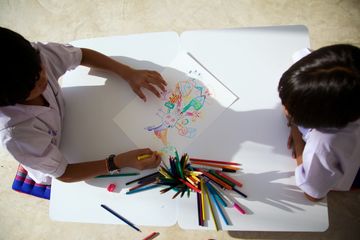 Two children seen from above drawing on one shared sheet, with the words Ping Pong Painting added over the photograph.