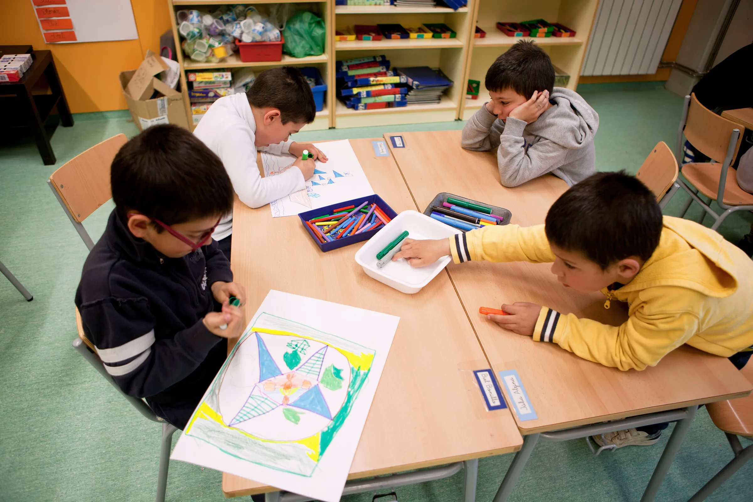 Children drawing around a table in a classroom with shelves of materials.