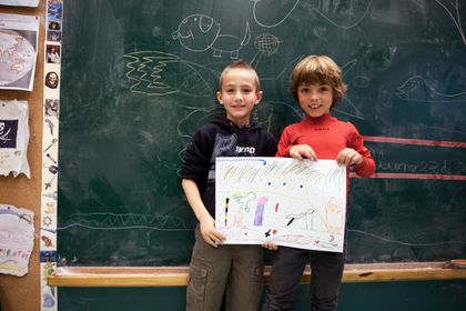 Two children in front of a chalkboard holding up a coloured drawing.