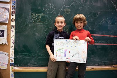 Two children in front of a chalkboard holding up a coloured drawing.