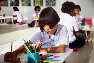 A child drawing at a low table beside a pot of coloured pencils, other children drawing behind.
