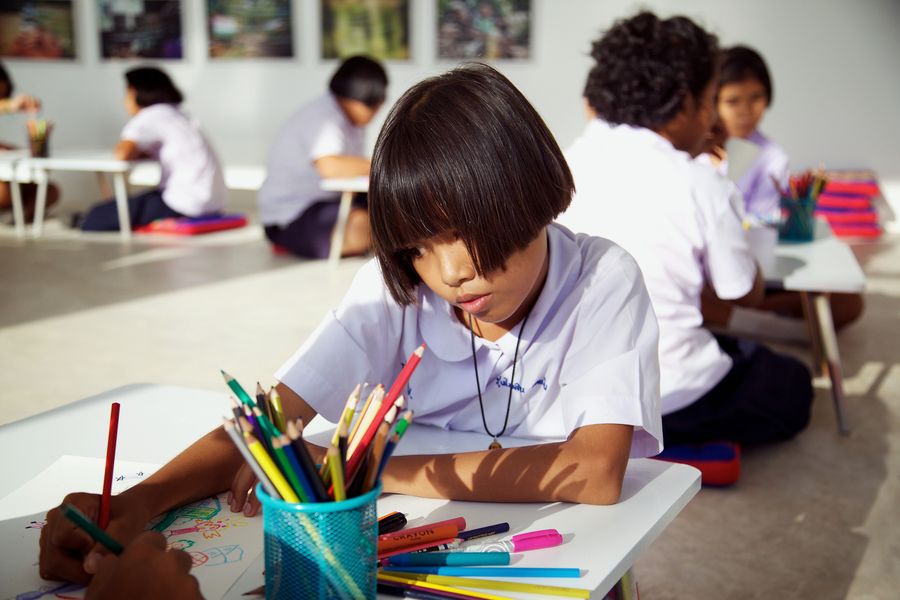A child drawing at a low table beside a pot of coloured pencils, other children drawing behind.