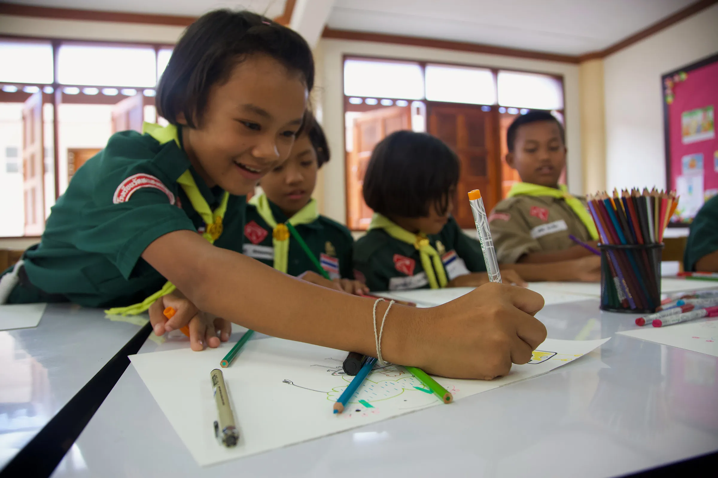 Children in green uniforms drawing at a long white table.