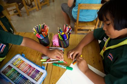 Two children drawing on one sheet at a wooden desk, with pots of felt pens between them.