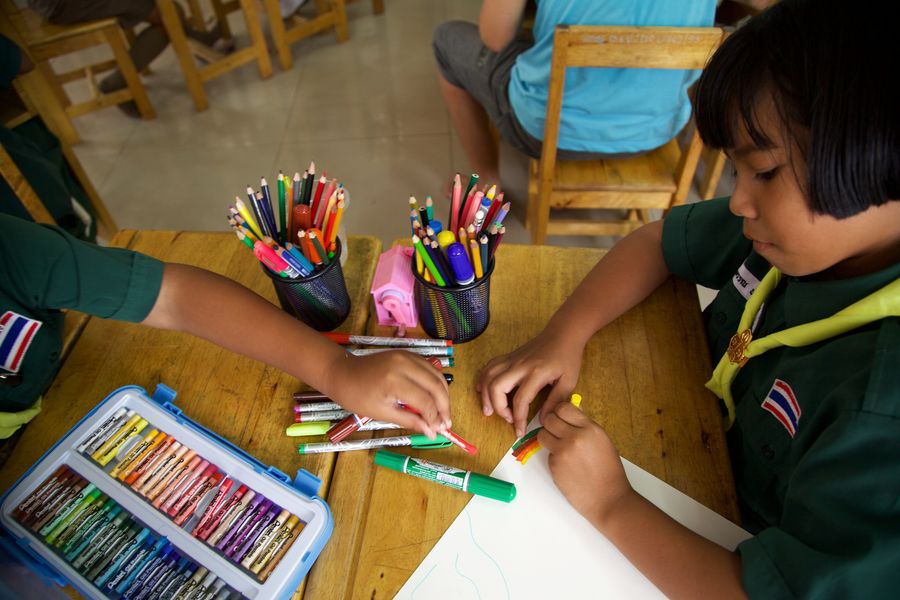 Two children drawing on one sheet at a wooden desk, with pots of felt pens between them.
