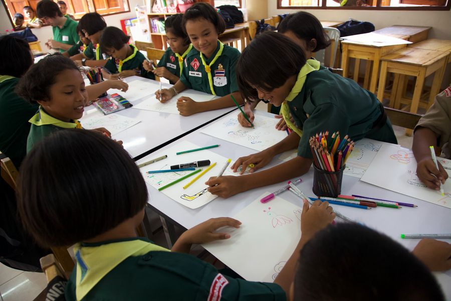 A classroom of children in green uniforms drawing on shared sheets at pushed-together desks.