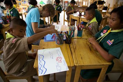 Children and an adult drawing at wooden desks; one sheet shows blue waves and red marks.
