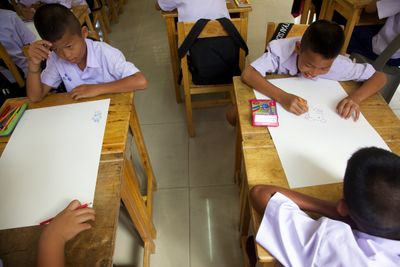 Children in white shirts drawing on large sheets at wooden desks.