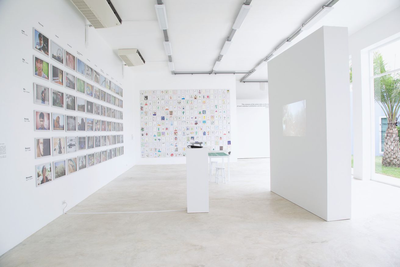 A white gallery room: a wall hung with rows of photographs on the left and, beyond a free-standing white wall, a wall of children's drawings in rows.