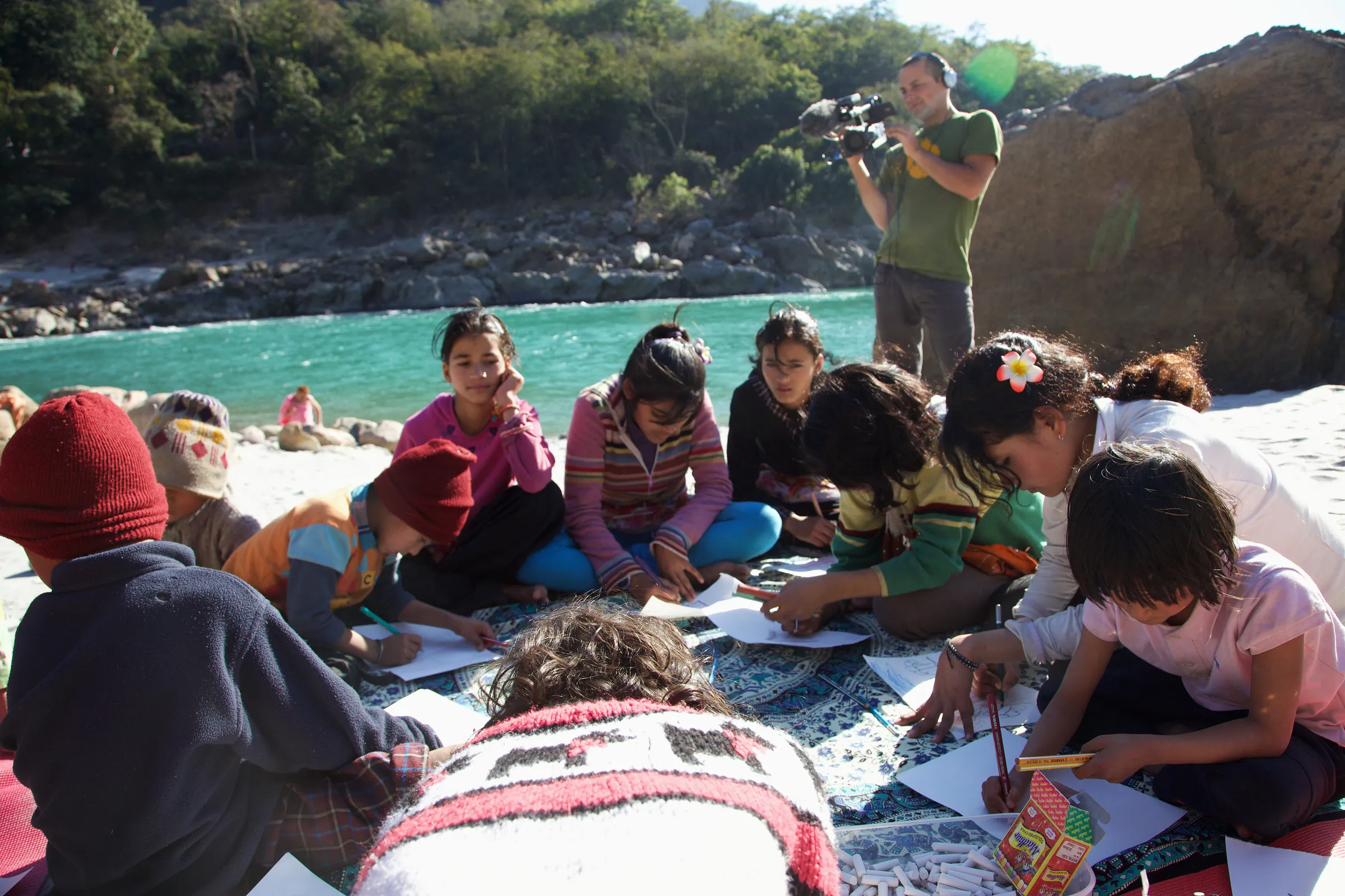Children drawing on sheets spread over a patterned cloth by a river, with a camera operator behind them.
