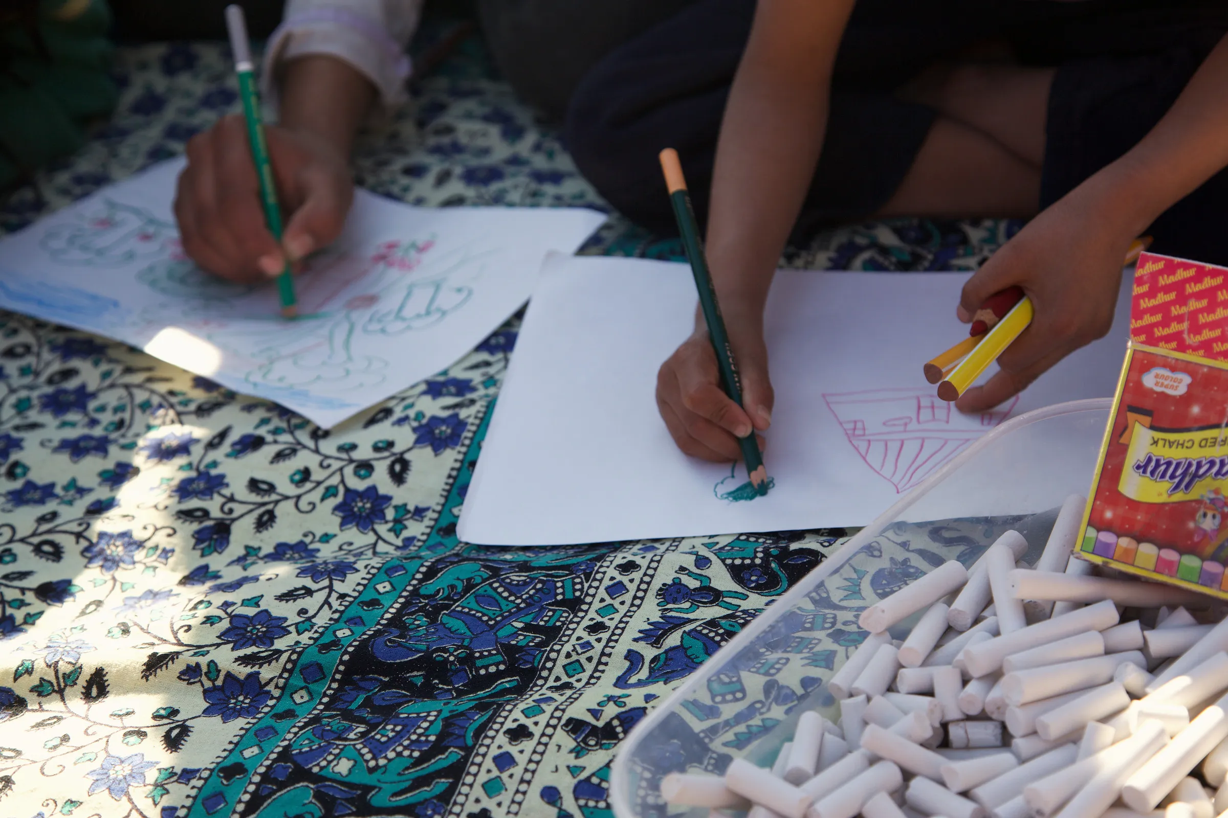 Hands drawing on paper on a patterned cloth, with sticks of chalk in a tray.