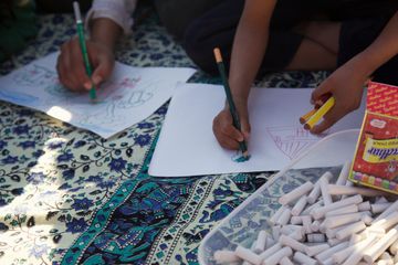 Hands drawing on paper on a patterned cloth, with sticks of chalk in a tray.