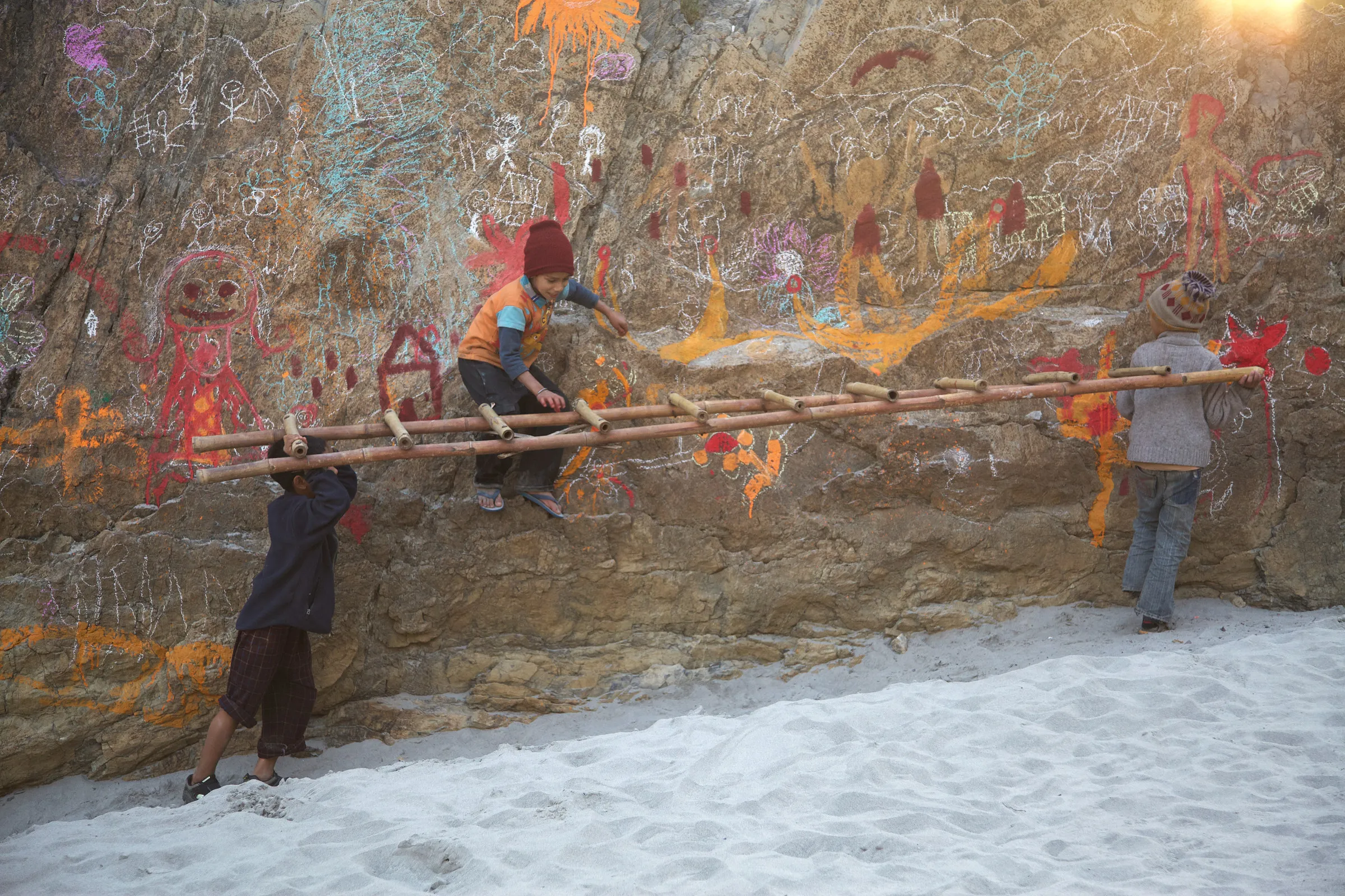 Children carrying a long ladder past a rock covered with chalk drawings.