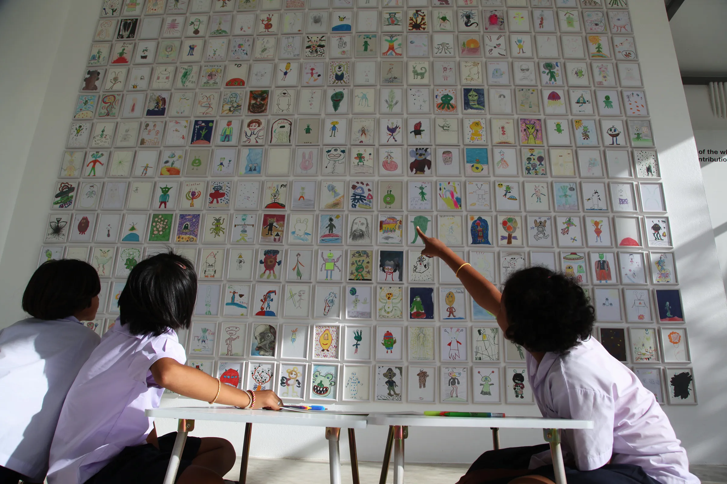 Children in front of a gallery wall covered with hundreds of alien drawings, one pointing at a drawing.