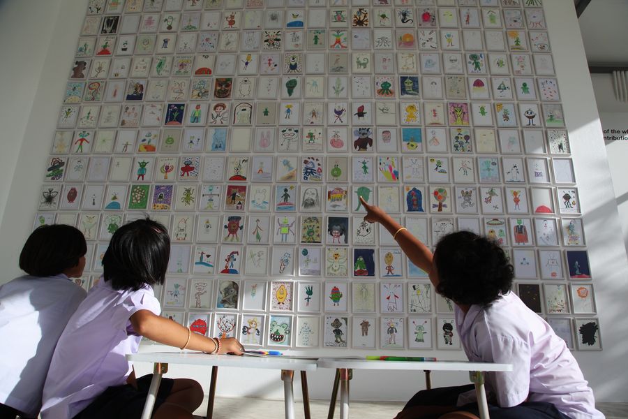 Children in front of a gallery wall covered with hundreds of alien drawings, one pointing at a drawing.