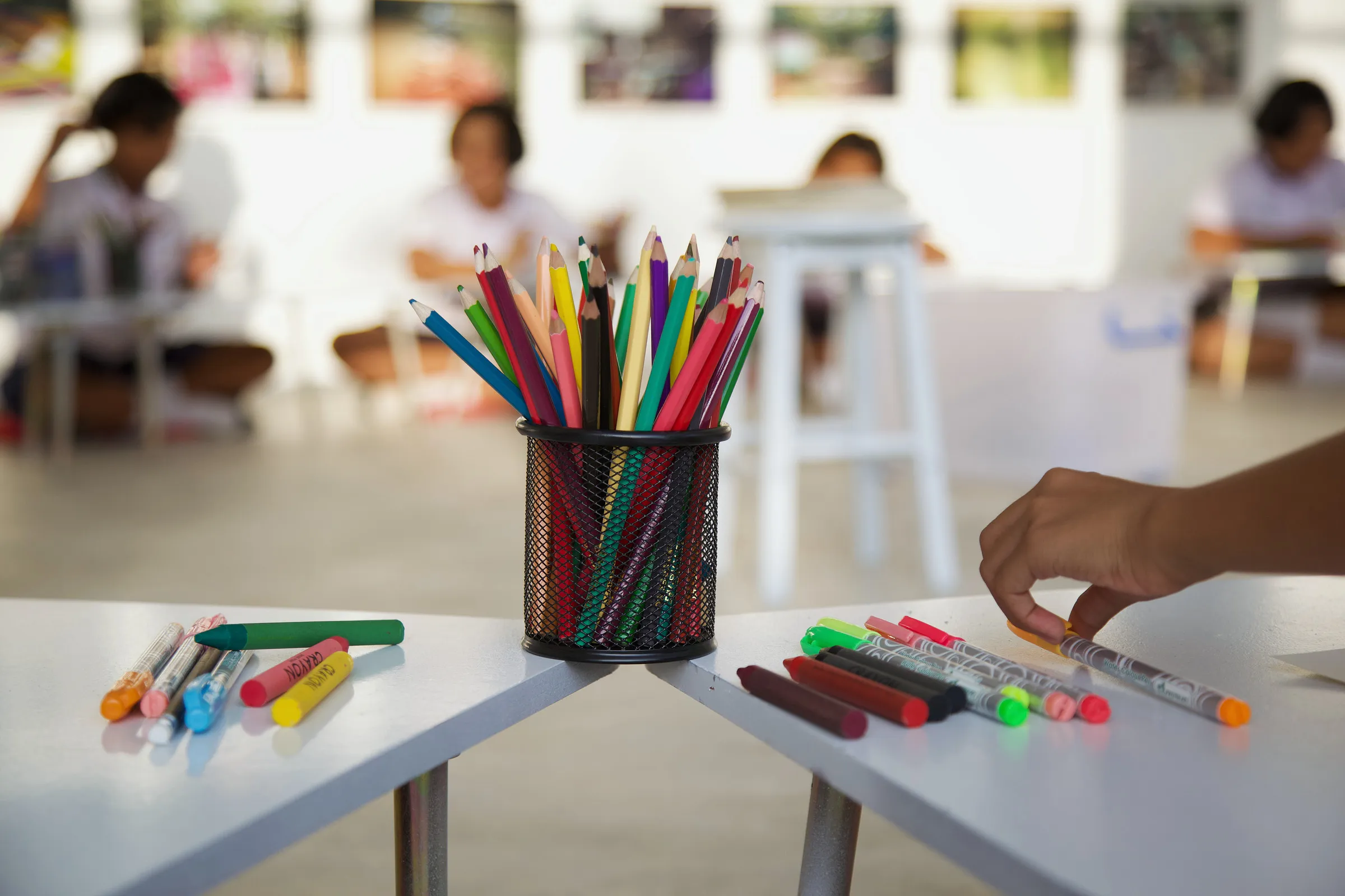 A pot of coloured pencils and scattered felt pens on a white table, with children drawing behind.