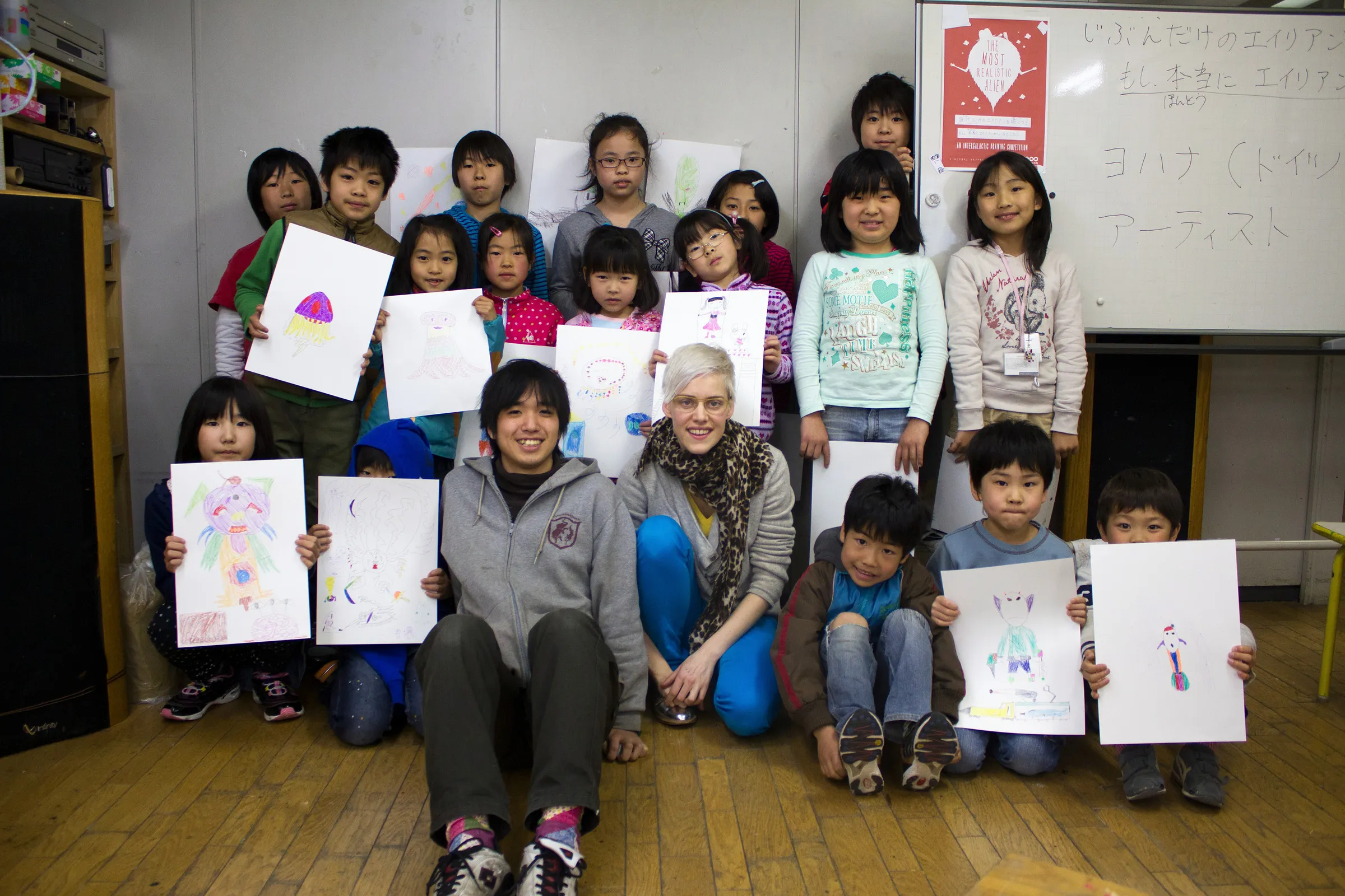 A group of children and two adults holding up alien drawings in a classroom.