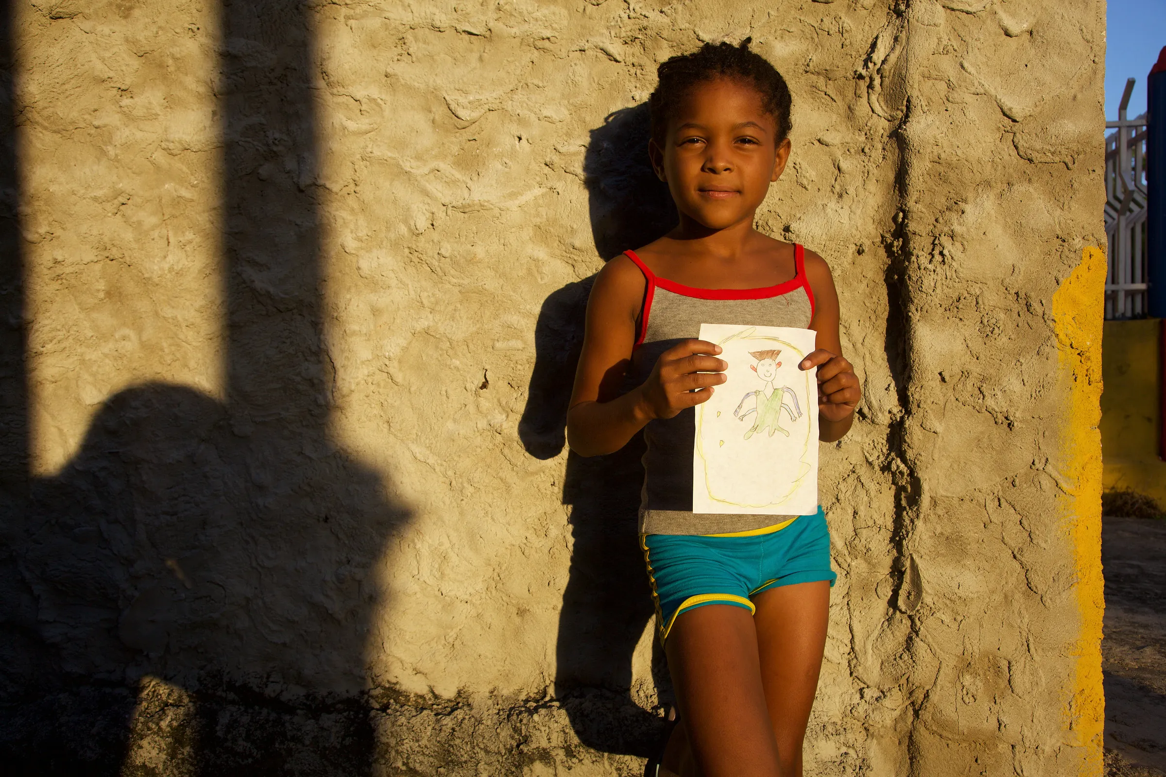 A child holding a small drawing in front of a sunlit wall.