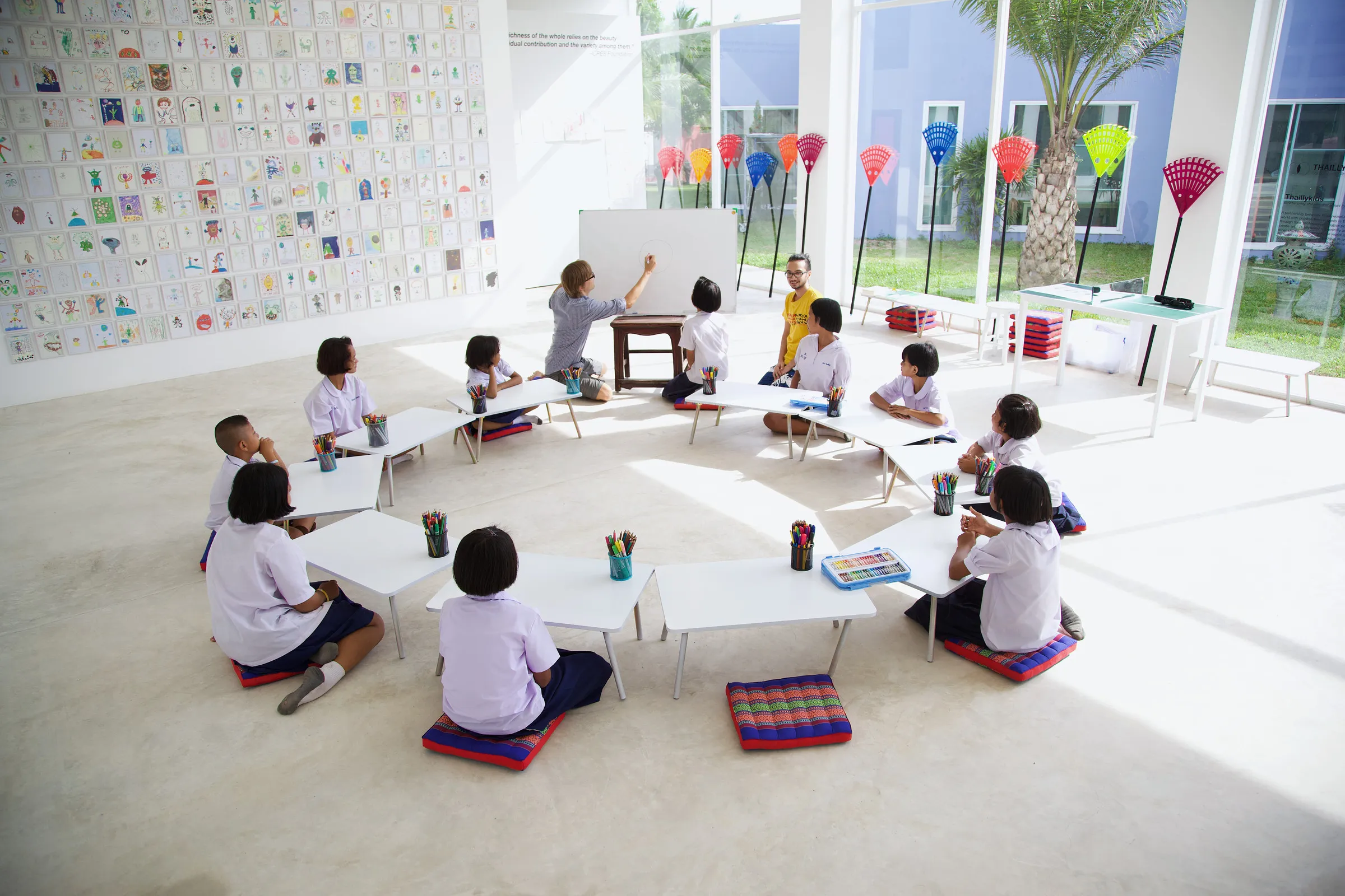 A bright room with children drawing at low white tables and alien drawings covering the back wall.