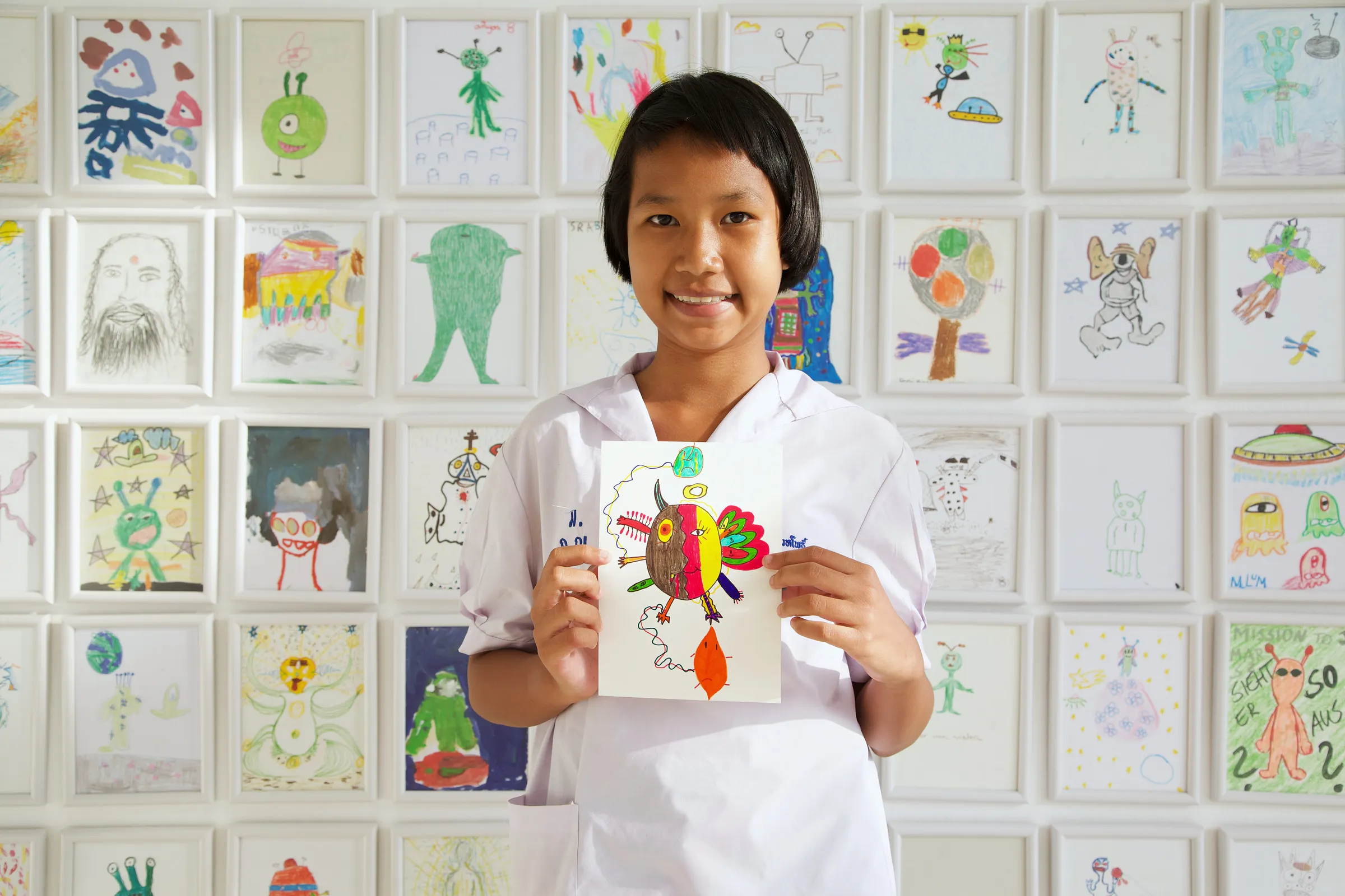 A child holding an alien drawing in front of a wall of framed alien drawings.